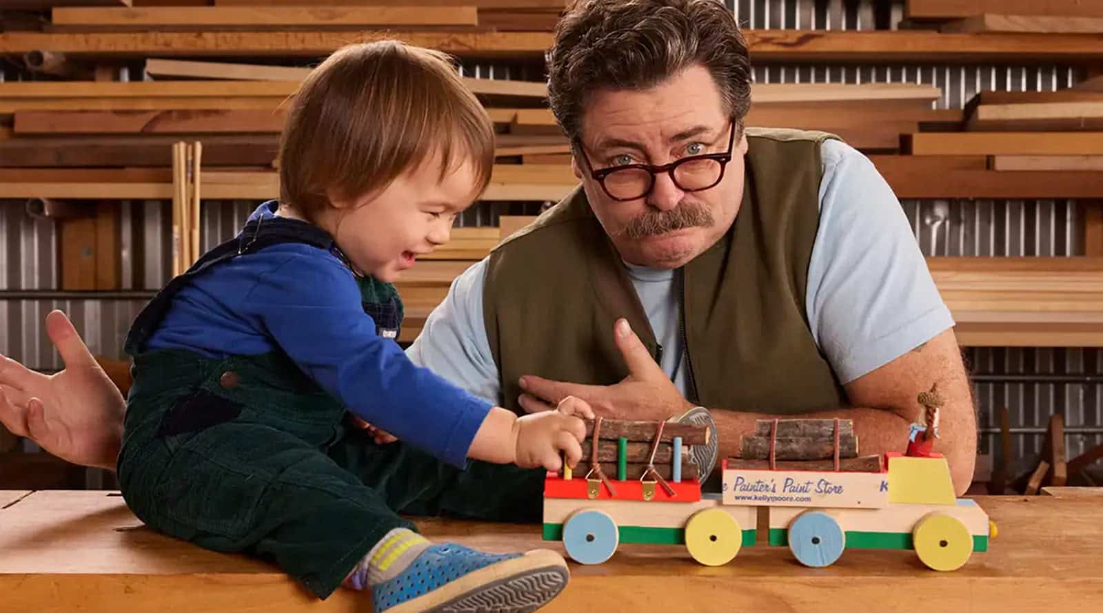 Nick Offerman posing with child and wooden toy from his book "Little Woodchucks: Offerman Woodshop's Guide to Tools and Tomfoolery".